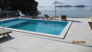 En rektangulär pool med en kakelbeklädd poolkant, två solstolar, ett bord och ett stort parasoll. Ett metallräcke omger poolen, och ett träd syns till vänster. I bakgrunden syns en vattenyta och ett avlägset landområde.