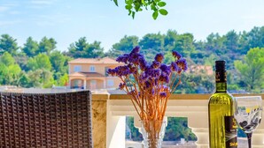 Une table avec une bouteille et un verre, un vase rempli de fleurs violettes et une chaise en osier.