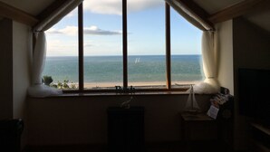 Ein Zimmer mit großem Fenster zum Strand, einem kleinen Tisch mit einem Modellschiff und einem Mülleimer.