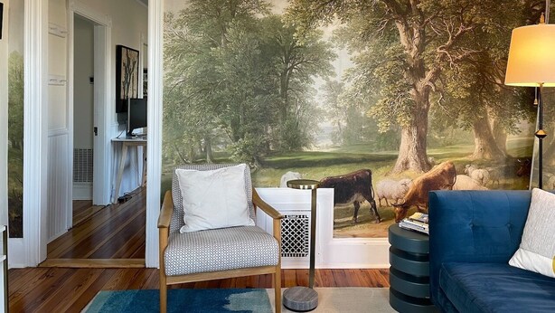 Een kamer met een muurschildering van een boslandschap, een blauwe bank, een gestreepe fauteuil en een kleine tafel met boeken.