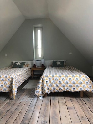 Um quarto de sótão pequeno e acolhedor, com duas camas simples, chão de madeira, uma janela e uma mesinha.