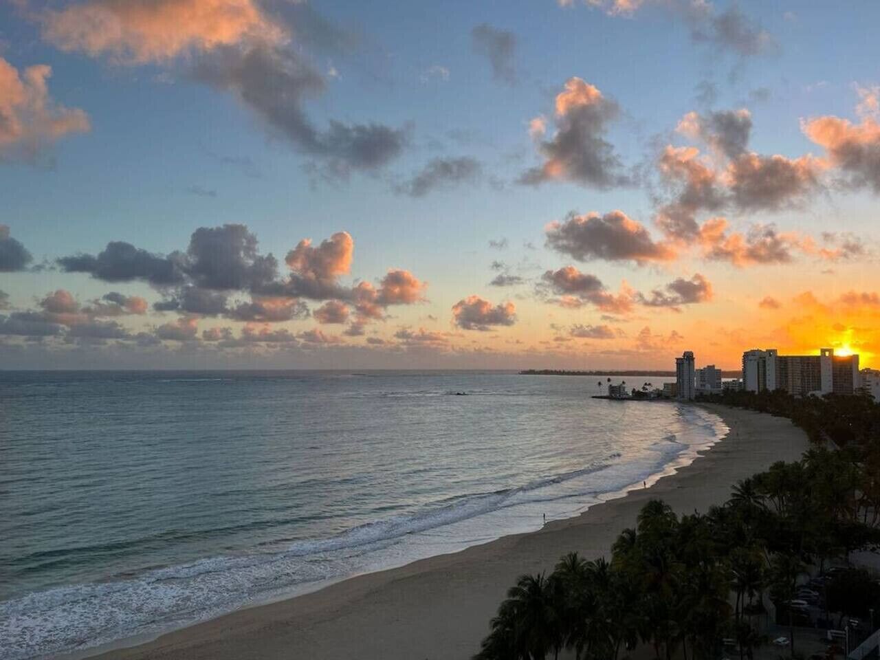 Ein Strand mit Sonnenuntergang, Meer und Gebäuden im Hintergrund.