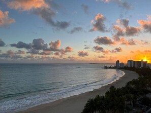A beach with a sunset, ocean, and buildings in the background.