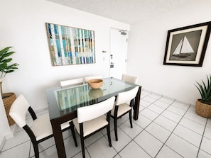 Uma sala de jantar com mesa de vidro, cadeiras brancas, cesto de vime e quadros nas paredes.