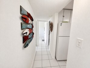 Un pasillo con piso de baldosas blancas, una pared blanca con un cuadro decorativo de peces y un refrigerador blanco a la derecha.