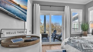 Une chambre avec un lit, un panneau indiquant « Bonjour », un plateau avec des télécommandes, une fenêtre avec des rideaux et une vue sur une terrasse.