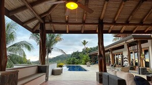 Área externa coberta com piscina, assentos e vista para palmeiras e colinas.