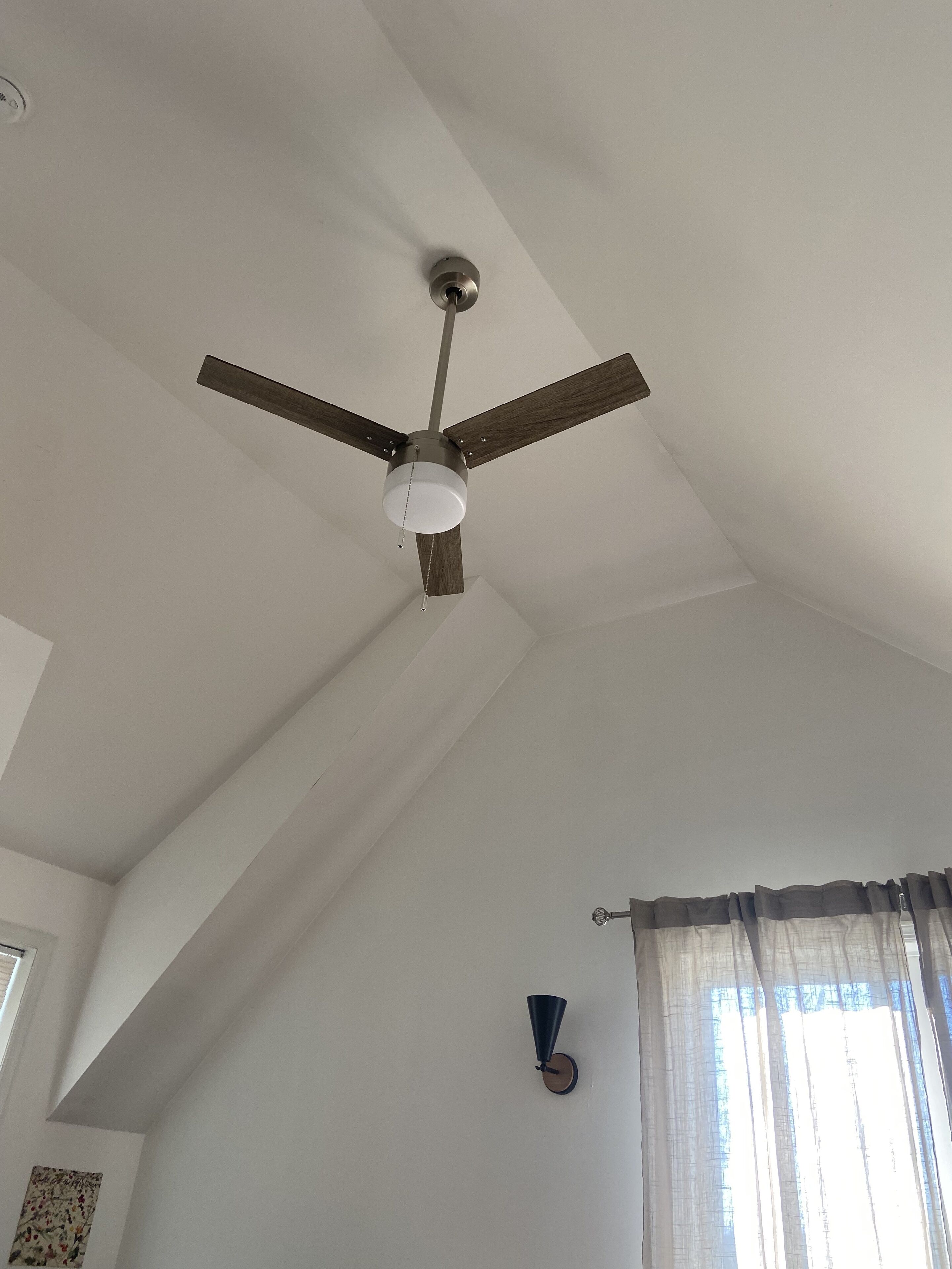 A ceiling fan with three blades and a light fixture in a room with white walls and a window with curtains.