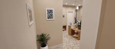 A hallway with beige walls, a potted plant, and a tiled floor. There are framed pictures on the walls and a door leading to a room with a mirror and a small table with a vase.
