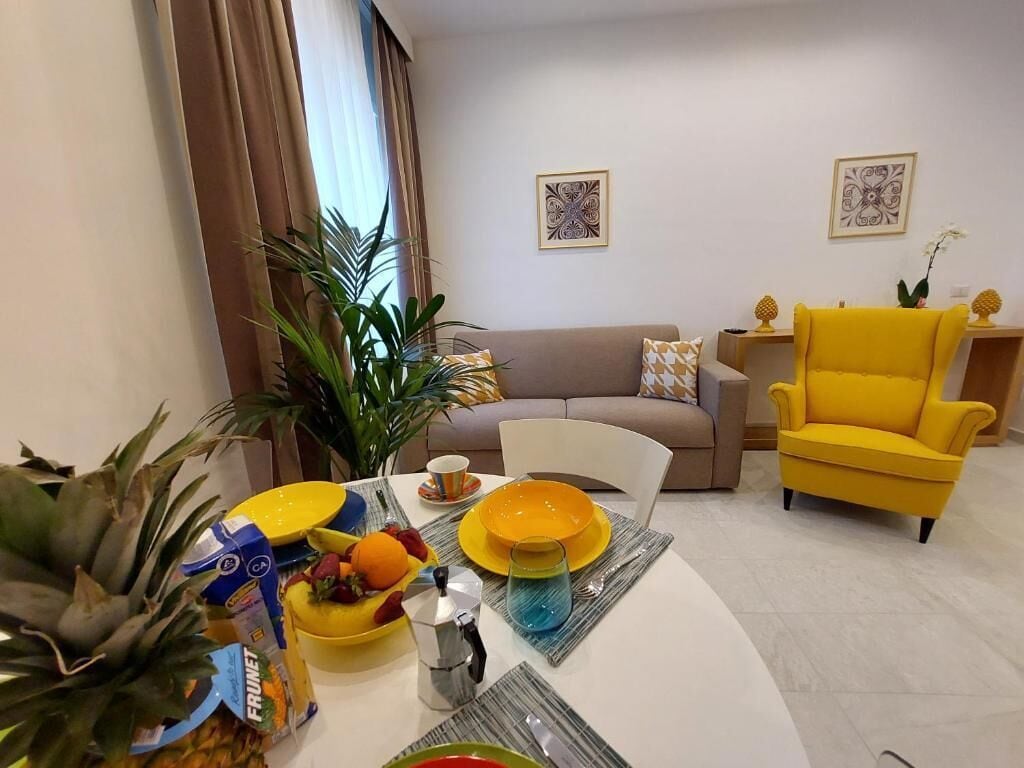Un salon avec un canapé, un fauteuil jaune et une table à manger sur laquelle on trouve des fruits et des boissons.