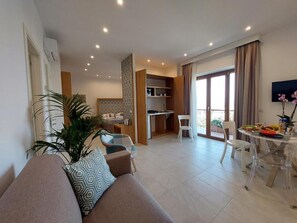 A modern living room with a sofa, a dining area with a table and chairs, a kitchen area, and a large window with curtains. - MISTRAL LUXURY SUITES (Sorrento)