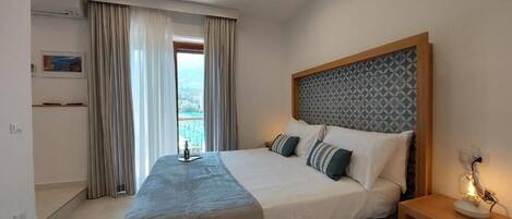 A modern bedroom with a large bed, a bedside table with a lamp, and a view of the outdoors through a window.