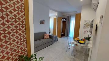 Una sala de estar moderna con un sofá gris, una mesa de comedor con sillas y una cocina que incluye un refrigerador y un lavamanos.