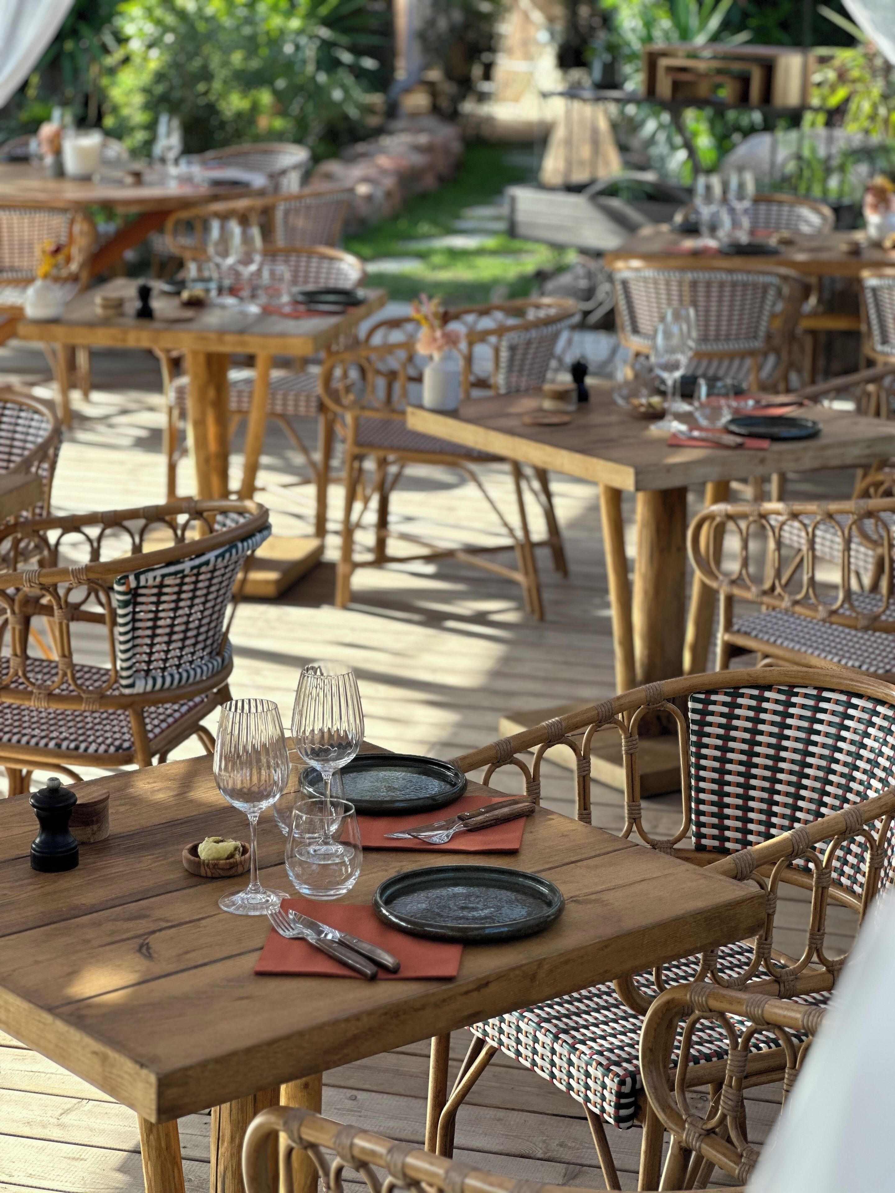Un espace de restauration en plein air, avec des chaises et des tables en osier, chaque table étant dressée avec des assiettes, des couverts et des verres à vin.