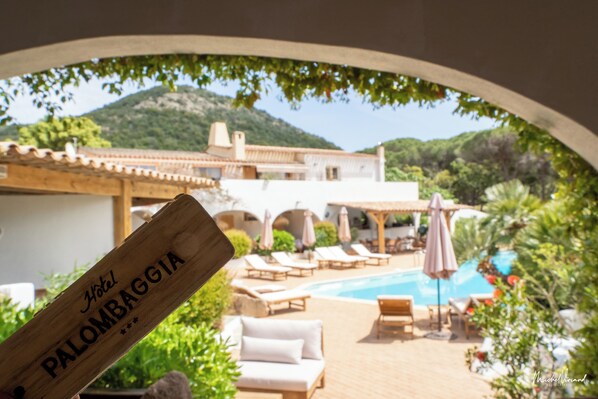 Uma placa de madeira com a inscrição 'Hotel Palombaggia', área de piscina com espreguiçadeiras e guarda-sóis, e montanhas ao fundo.