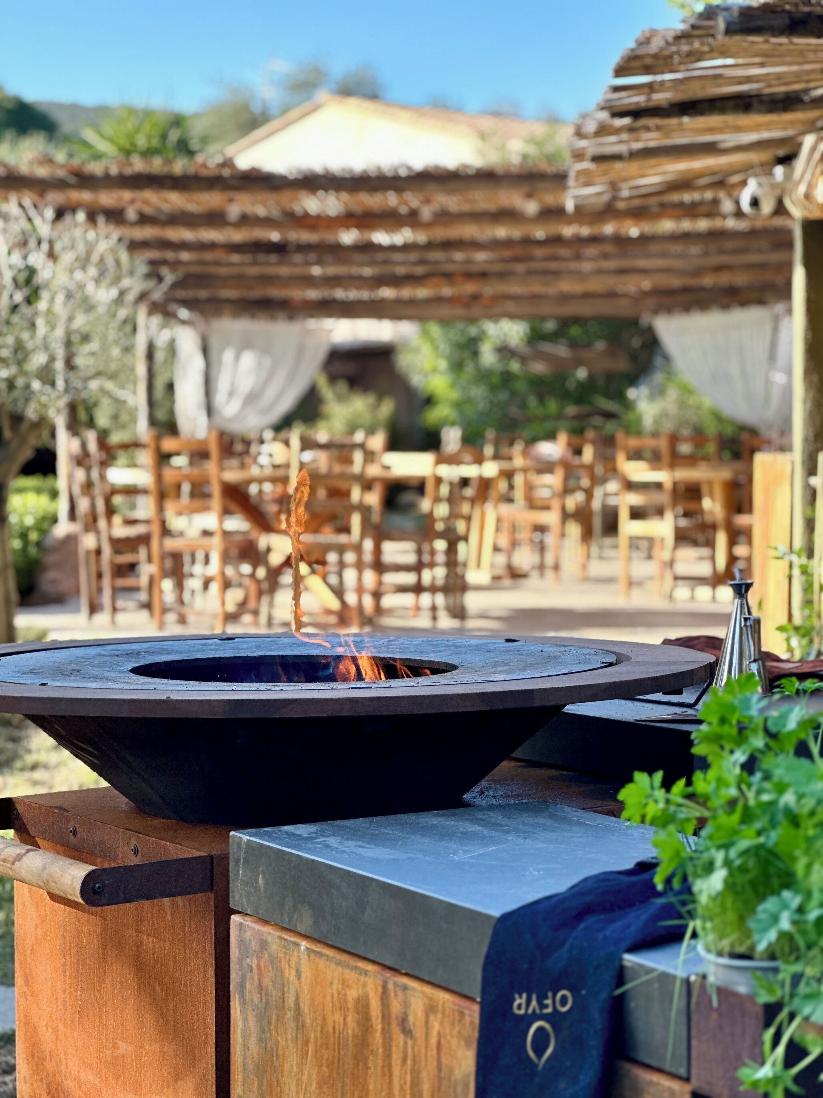 An outdoor dining area with a fire pit in the foreground and wooden tables and chairs in the background.