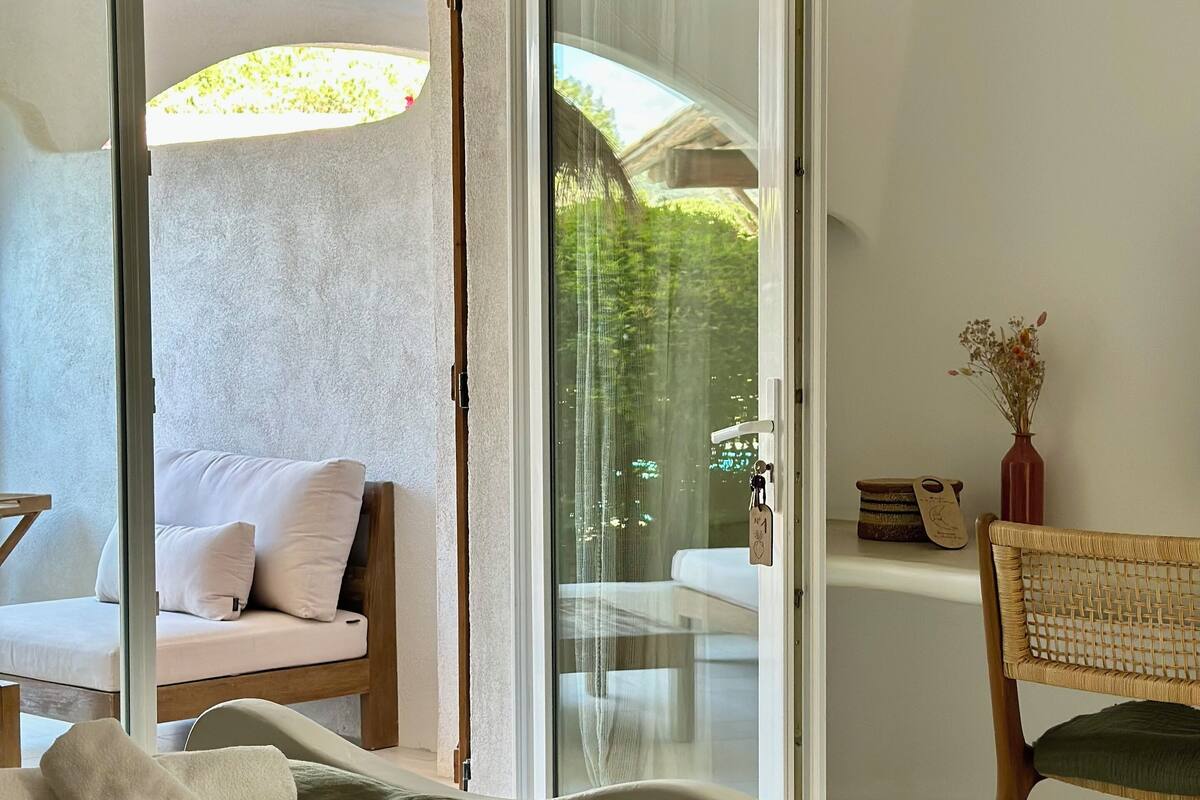 A modern bedroom with a large bed, a sliding glass door leading to a balcony, a wicker chair, and a small table with a vase.