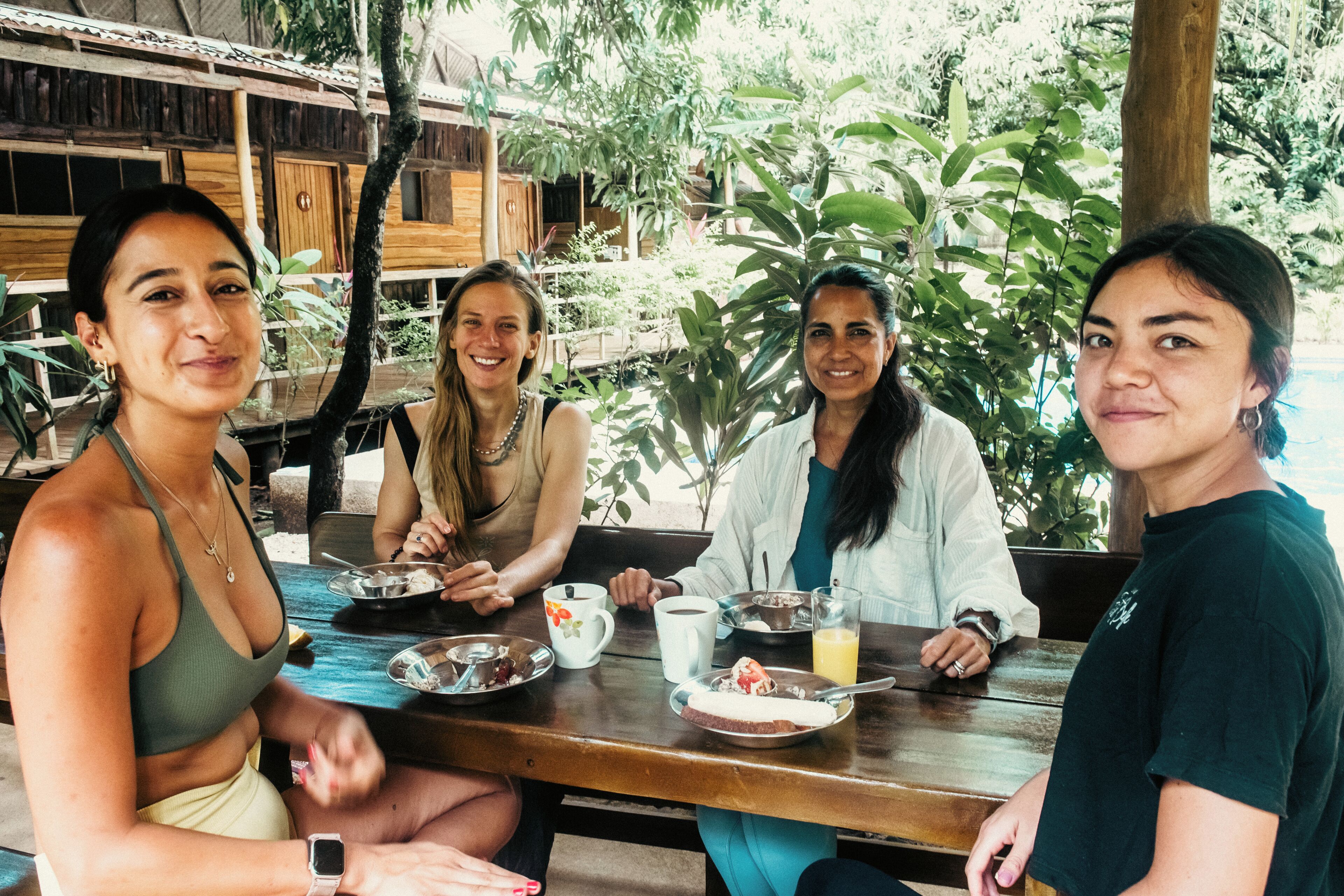 Four people seated at an outdoor table with plates of food, cups, and a glass of juice.