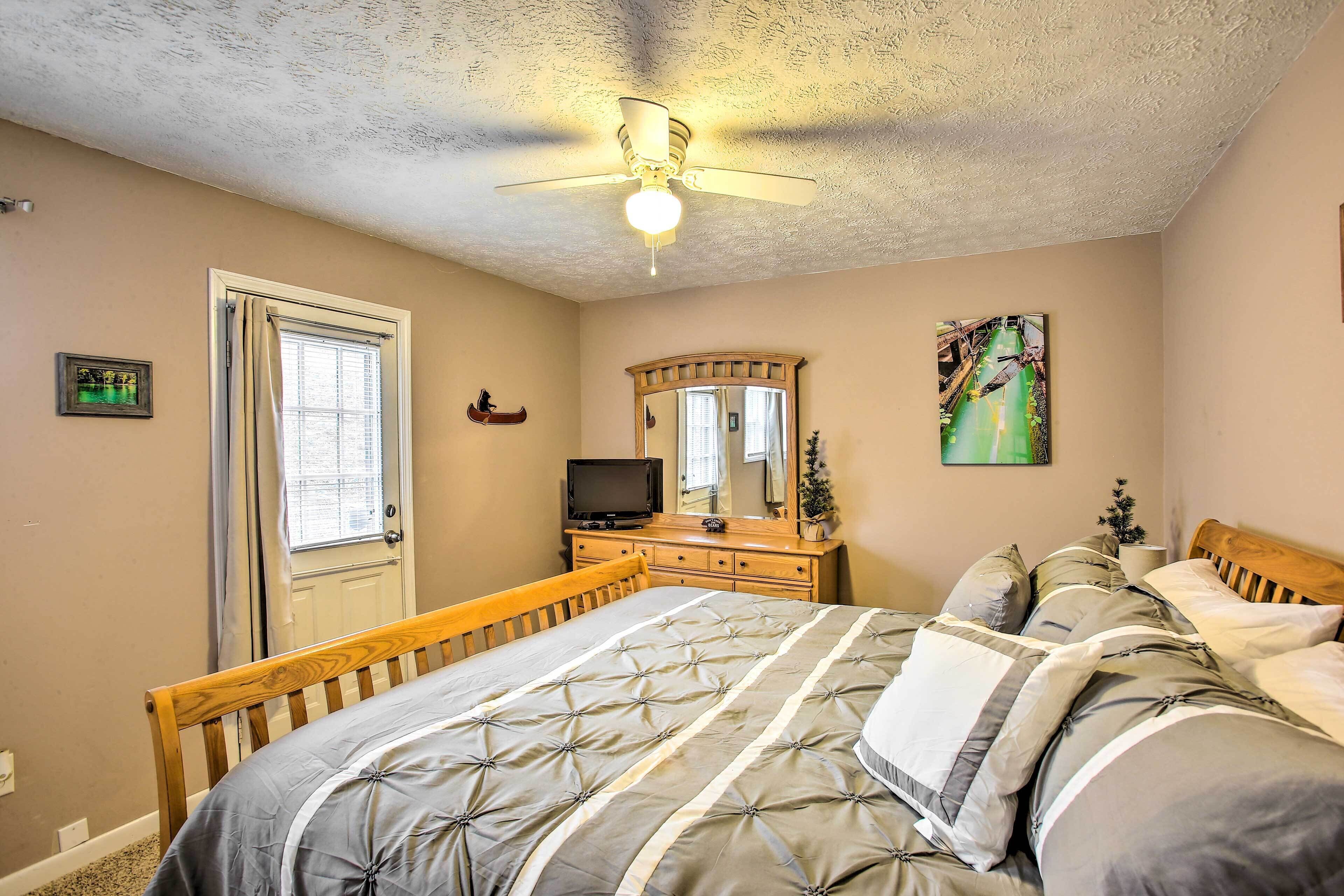 A bedroom with a bed, wooden headboard, a ceiling fan, a dresser, a television, a mirror, and a window with curtains.