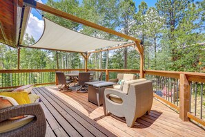 Relax on the deck surrounded by a forest, with comfortable wicker sofas and chairs, a dining table, and a large umbrella providing shade.