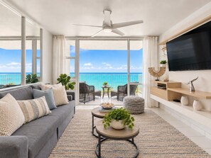 A spacious living room with a large window overlooking the ocean, a ceiling fan, a flat-screen TV, and a comfortable seating area. - Beachfront 2b/2b  Walk 2 Eat  Isla Verde Beach (Carolina)