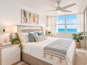 Un dormitorio con cama, mesitas de noche, ventilador de techo, ventana con vista al océano y un cuadro de piña en la pared.