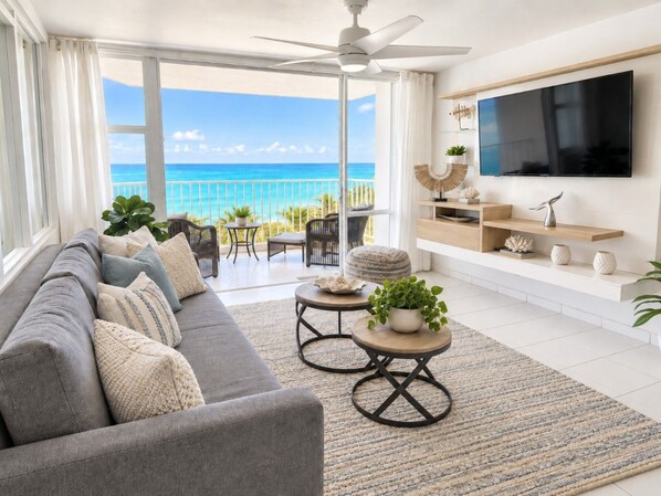 A modern living room with a grey sofa, two round tables, a flat-screen TV, and a view of the ocean. - Beachfront 2b/2b  Walk 2 Eat  Isla Verde Beach (Carolina)