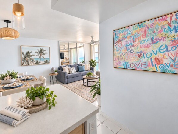 A modern living room with a dining area, a sofa, and a coffee table. There is a large abstract painting on the wall and a window providing natural light. - Beachfront 2b/2b  Walk 2 Eat  Isla Verde Beach (Carolina)