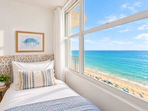 A bedroom with a large window overlooking a beach, a bed with patterned bedding, and a framed picture on the wall. - Beachfront 2b/2b  Walk 2 Eat  Isla Verde Beach (Carolina)