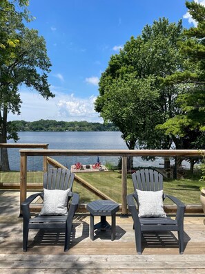 Une terrasse en bois avec deux fauteuils Adirondack et une petite table, d’où l’on a vue sur un lac, avec un quai et des chaises visibles au loin.