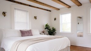 A bedroom with a large bed, wooden beams, white walls, and two windows with blinds.