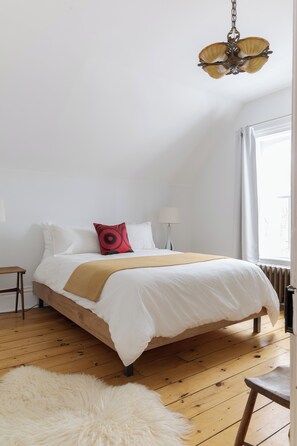 A bedroom with a large bed, a wooden floor, a fluffy rug, a red pillow, a lamp, and a ceiling light fixture.