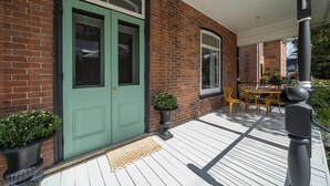 Un bâtiment en briques avec une porte et des fenêtres vertes, une terrasse en bois avec des meubles d’extérieur et un poteau noir.