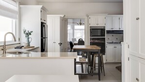 A modern kitchen with white cabinetry, a central island, and a dining area with a wooden table and chairs. - The Carrington | 5 Bed Victorian | Pool  and  Hot Tub (Prince Edward)