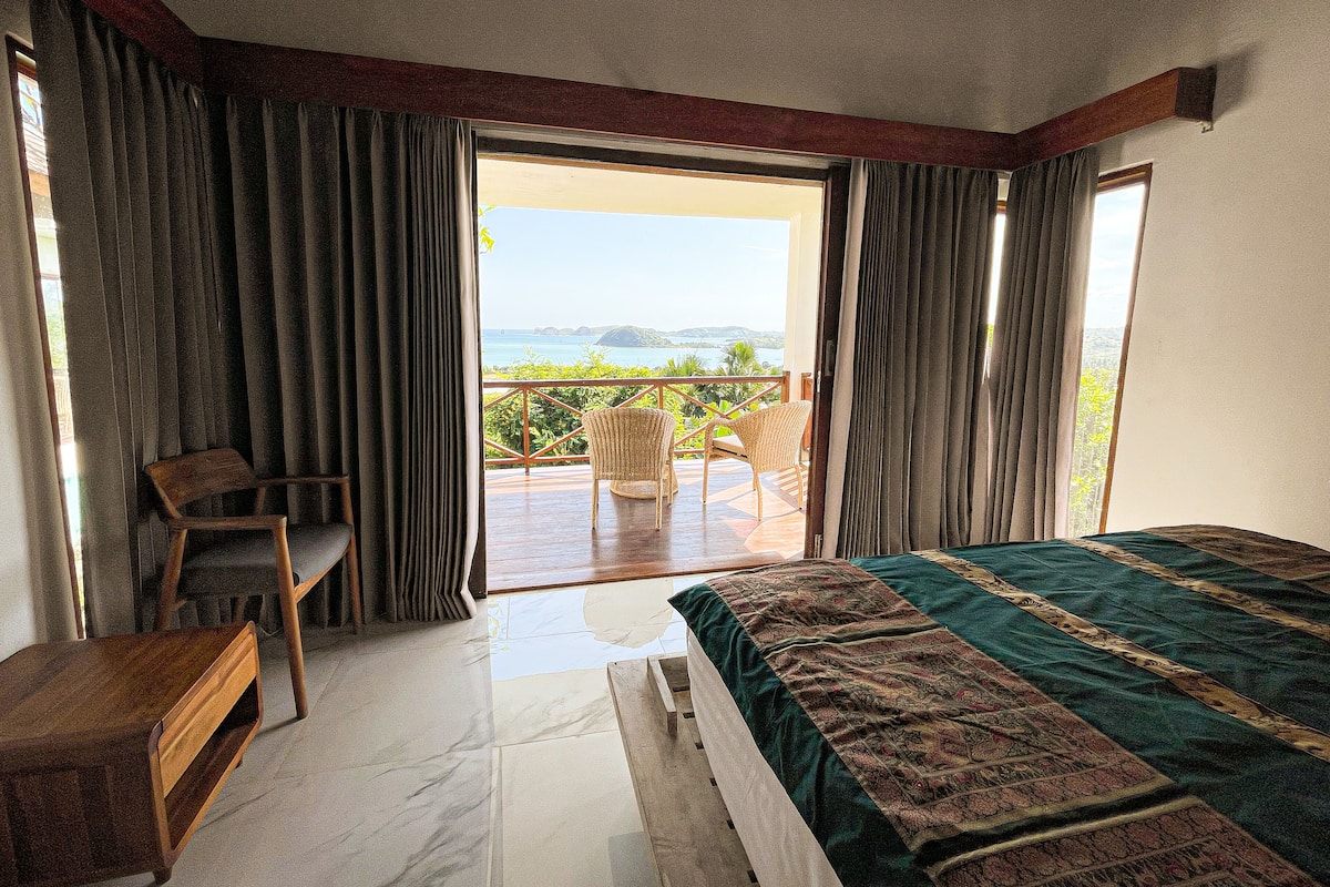 Quarto com uma cama grande, mesa de cabeceira de madeira, uma cadeira e vista para uma varanda com mesa e cadeiras.
