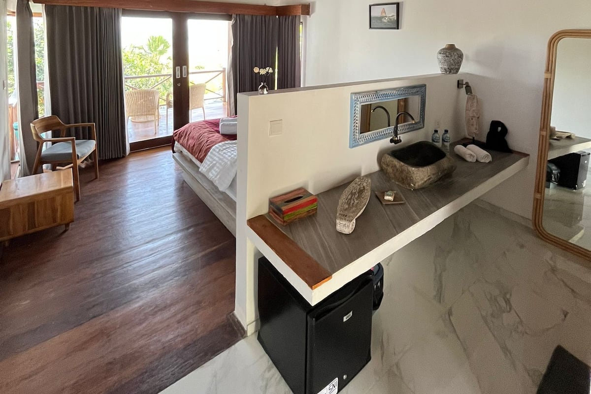 Quarto com cama, escrivaninha, espelho, pia e um pequeno eletrodoméstico preto.