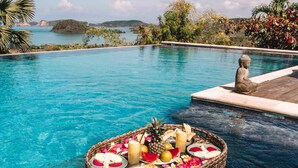 Un área de piscina tranquila con una cesta flotante que contiene comida y bebidas, rodeada de exuberante vegetación y una vista despejada del mar.