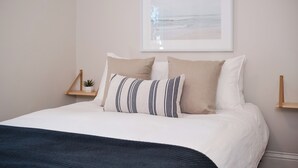 Una cama bien hecha con un almohadón a rayas, un almohadón liso y un almohadón color beige. A la izquierda de la cama, hay una pequeña planta sobre un estante de madera.