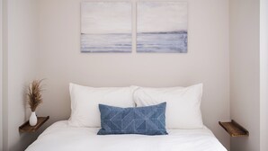 Een slaapkamer met een bed, witte kussens, een blauw kussen, een kunstwerk aan de muur en een klein houten plankje met een vaas.