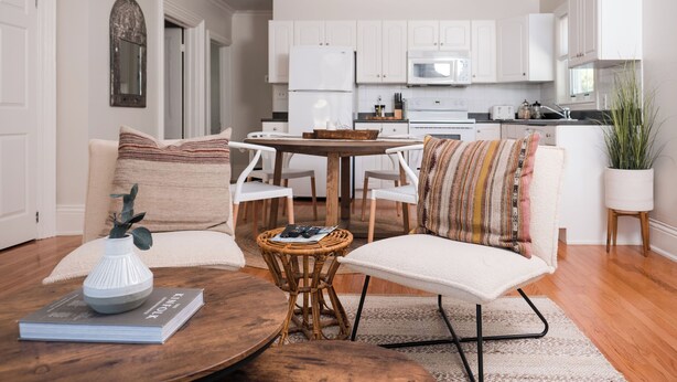 Een moderne woonkamer met een houten salontafel, een rieten bijzettafeltje en een witte bank met gekleurde kussens. In de achtergrond staat een eettafel met stoelen, en er is een keuken met witte kasten en apparatuur zichtbaar.