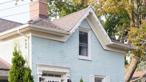 A light blue house with a white front porch, a chimney, and a small garden with various plants and flowers. - The Ferg Heritage Suite | Picton | 2 Bedrooms (Prince Edward)