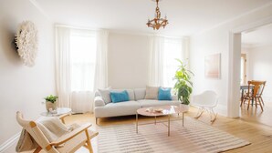 A spacious living room with a light-colored sofa, a coffee table, and a rocking chair. There is a large window with sheer curtains, a chandelier, and a potted plant. - The Ferg Heritage Suite | Picton | 2 Bedrooms (Prince Edward)