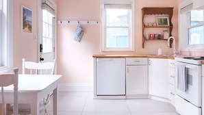 Una cocina con armarios blancos, lavavajillas, pileta y estufa. Hay una ventana con persianas, una puerta y un estante pequeño con frascos.