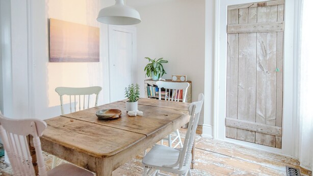 Un comedor con una mesa de madera, sillas blancas, una lámpara colgante, una planta en maceta y una estantería con libros.