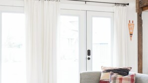 Ein Wohnzimmer mit grauem Sofa, einem Holztisch und einem Buch. Ein Schiebetürfenster mit weißen Vorhängen.