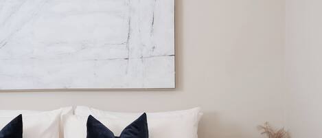 Un dormitorio con un cuadro abstracto grande, una cama con almohadas blancas y oscuras, una manta beige y un pequeño estante de madera con un florero.