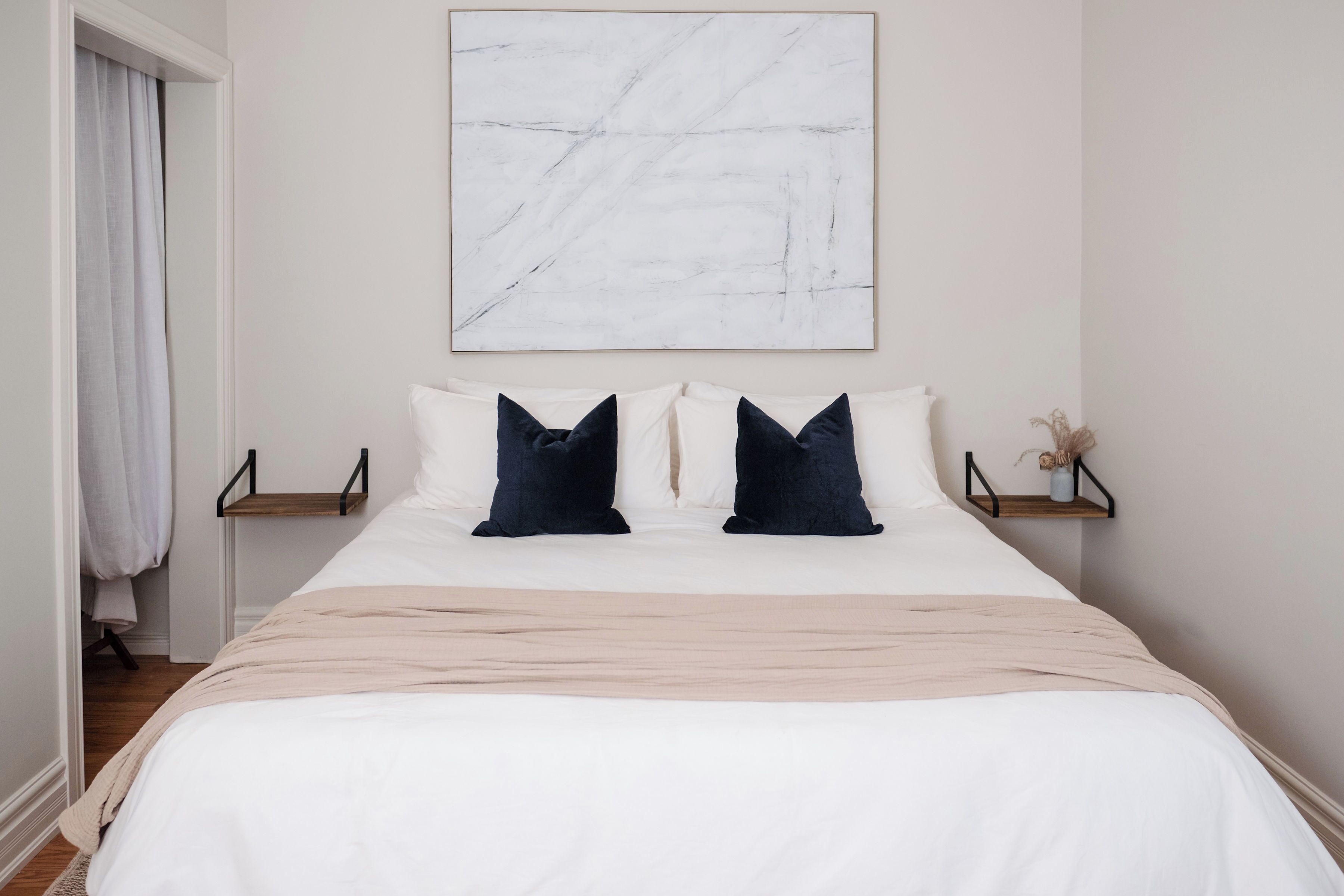 A neatly made bed with a white duvet, two dark pillows, and a light-colored throw. There is a marble-patterned artwork above the bed, and a small shelf with a vase of dried plants on the right side.