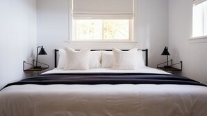 A bedroom with a large bed, two bedside tables with lamps, a window with blinds, and a view of trees outside. - The Wilolea | Milford | 5 Bedrooms | Waterfront (Prince Edward)