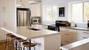 A modern kitchen with white cabinetry, stainless steel appliances, a central island with stools, and pendant lighting. - The Wilolea | Milford | 5 Bedrooms | Waterfront (Prince Edward)