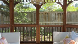 A covered outdoor seating area with two white sofas, a woven ceiling light, and a wooden fence backdrop. - The Addison | Modern Farmhouse | Pool  and  Hot Tub (Prince Edward)